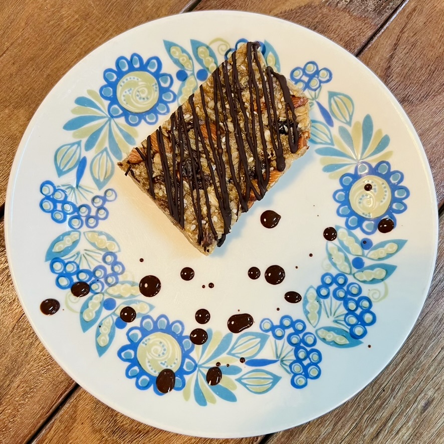 A rectangular muesli slice drizzled with dark chocolate sits on a white plate with blue and green floral patterns. Small droplets of chocolate are scattered on the plate, which rests on a wooden table.