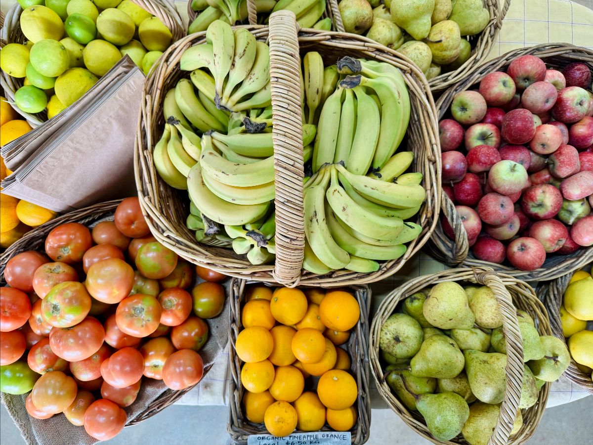 Overhead view of wicker baskets filled with green bananas in the center, surrounded by red apples, green pears, lemons/limes, oranges, and tomatoes on a checkered table. A stack of brown paper bags sits beside the citrus basket.