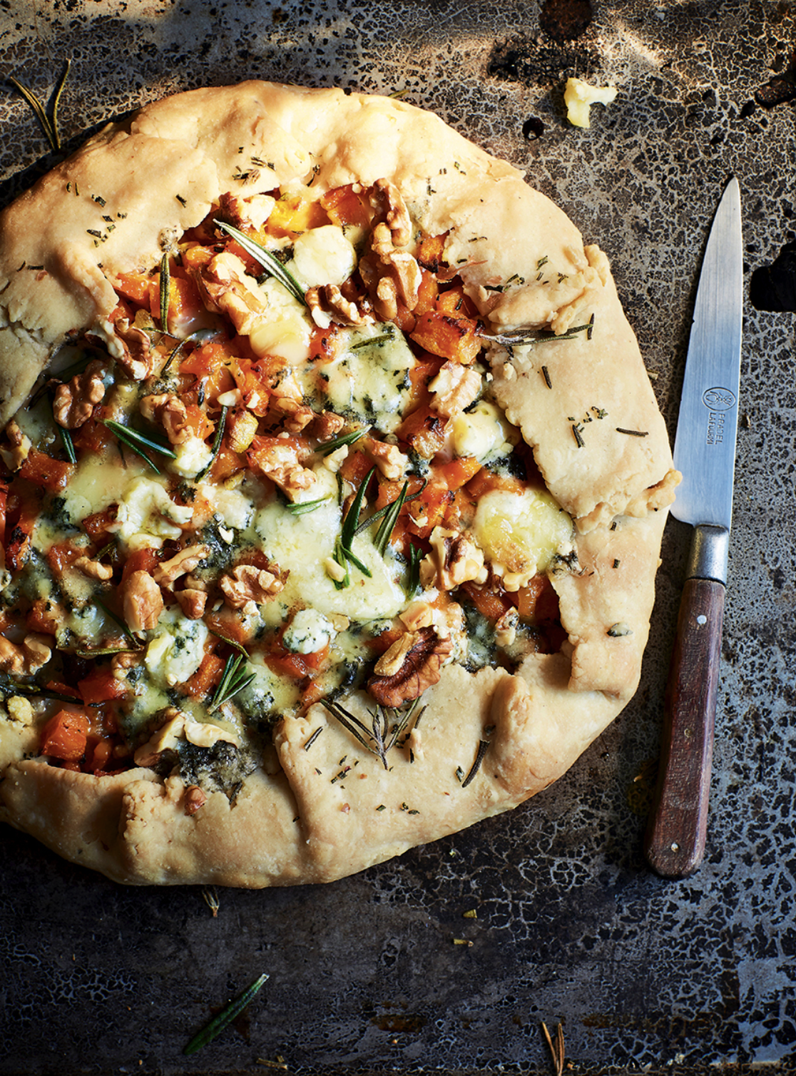 Rustic round galette with folded crust, topped with orange pumpkin pieces, melted blue cheese, walnuts, and rosemary on a dark metal baking tray. A knife with a wooden handle lies to the right.
