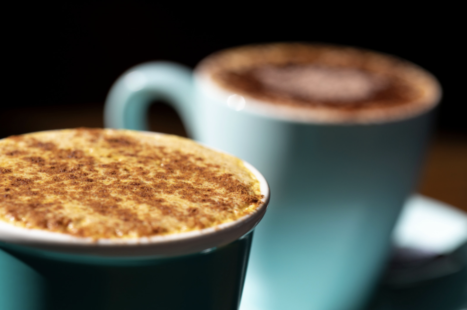 Close-up of two teal ceramic mugs filled with hot drinks; the front mug has a foamy top dusted with brown spice, while the second mug is blurred in the background. The mugs sit against a dark, out-of-focus backdrop.