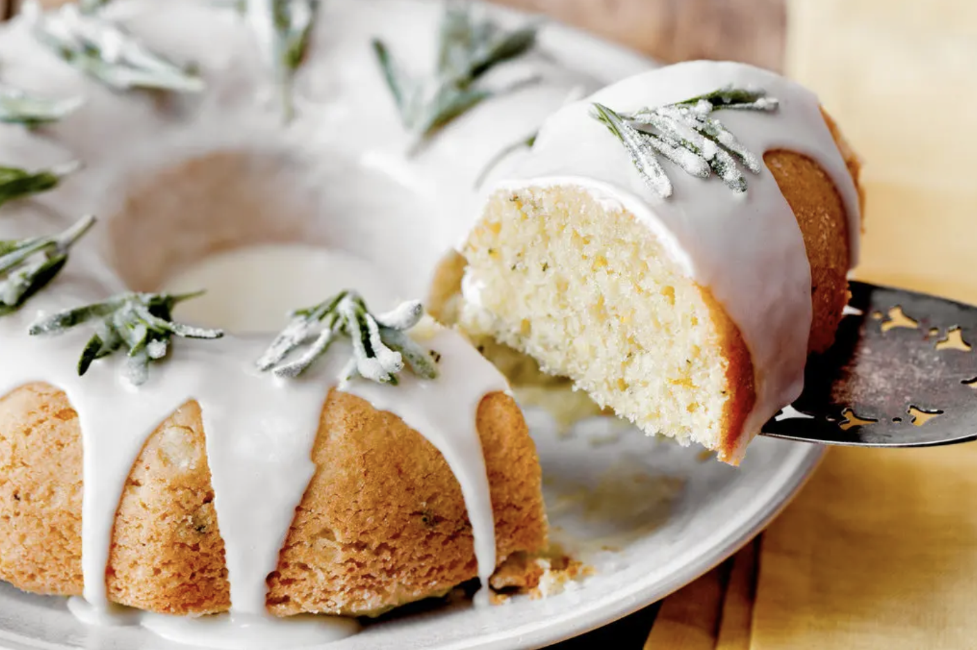 A glazed bundt cake on a plate, topped with sugared rosemary sprigs. A slice has been cut out and is being lifted on a spatula, showing the cake’s light crumb.