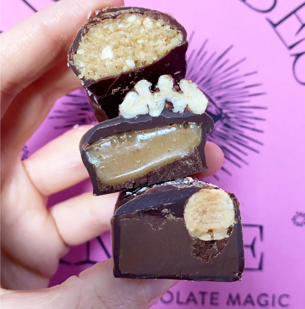Hand holding three stacked dark-chocolate candies, each cut open to show different fillings: a crumbly nut center, a caramel-like layer, and a whole hazelnut. Pink packaging with black line art is blurred in the background.