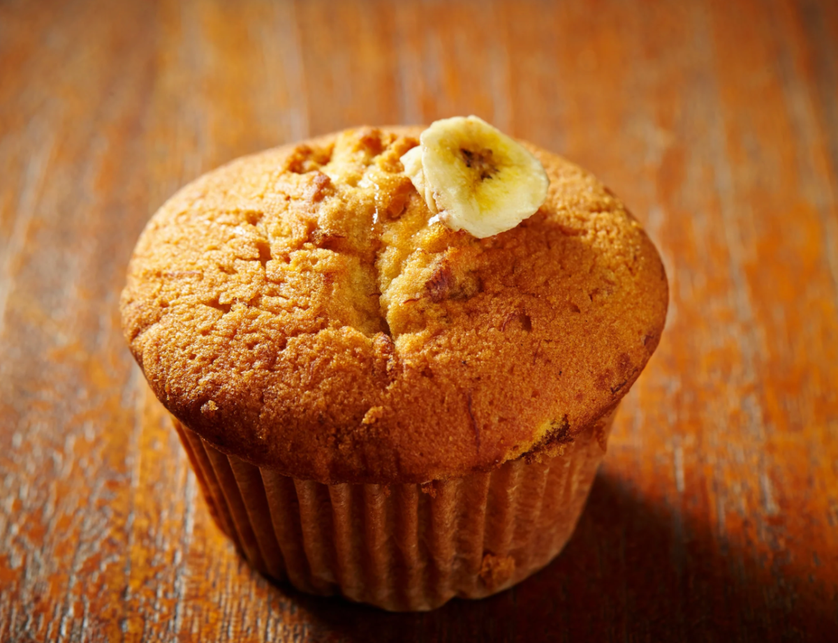Close-up of a banana muffin in a brown paper liner on a wooden table. A banana slice sits on top of the muffin.
