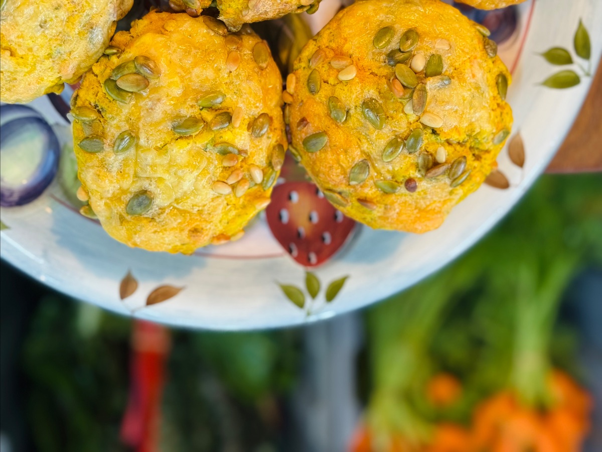 Close-up of two golden muffins topped with pumpkin seeds on a patterned plate. The background is blurred with green foliage and orange flowers.