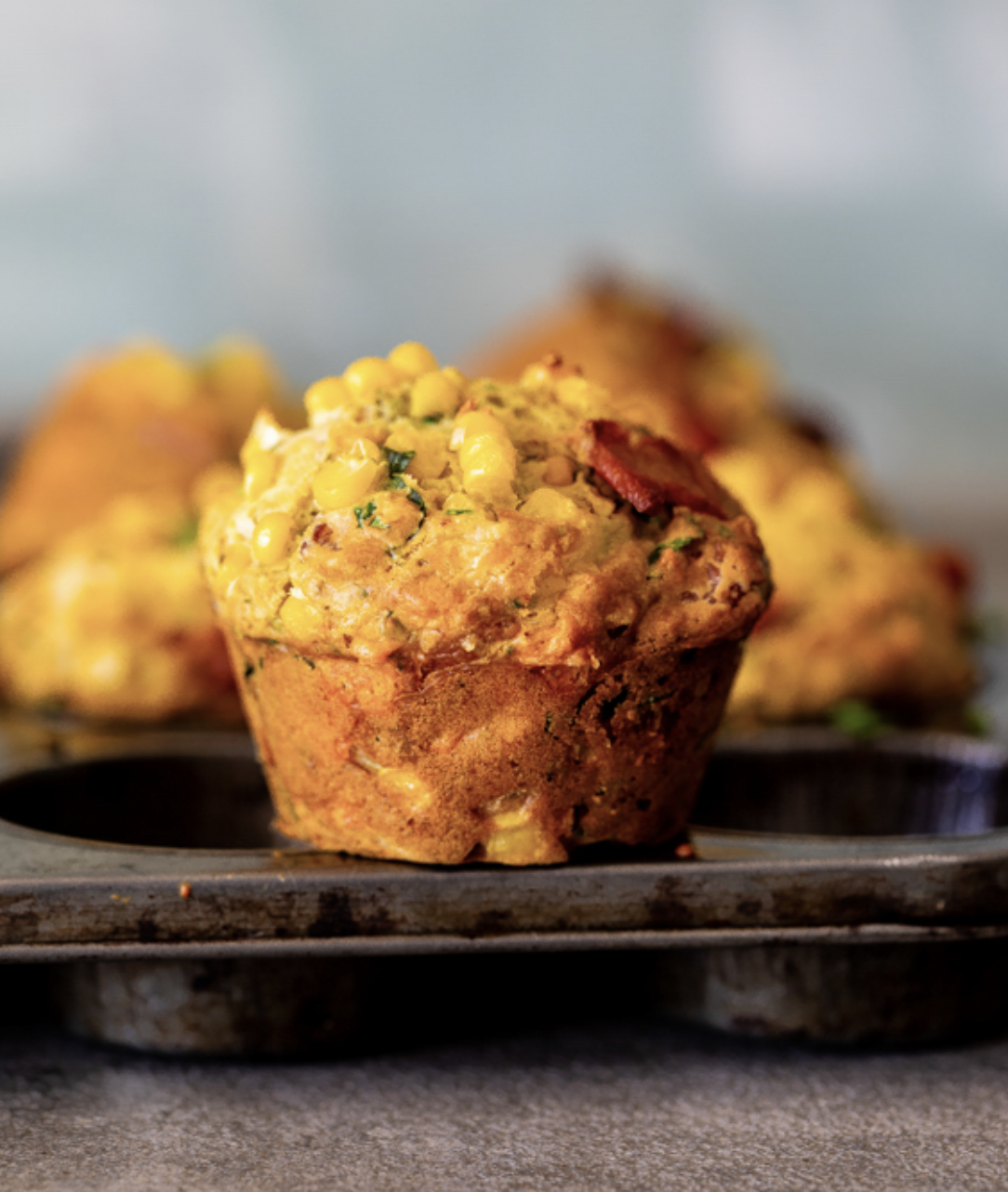 Close-up of a golden savoury muffin topped with corn kernels and herb flecks in a metal muffin tin. Several more muffins are visible out of focus in the background.