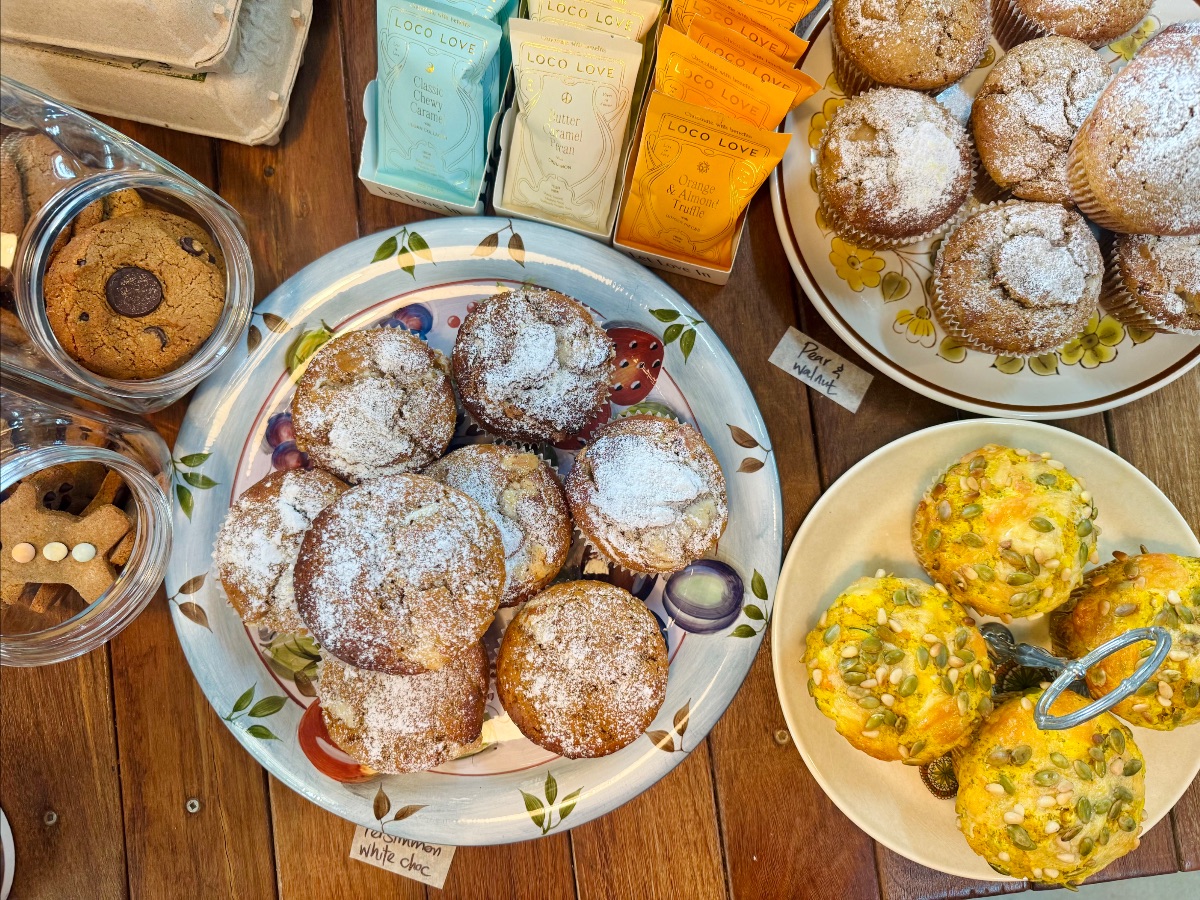 Overhead view of a wooden table with plates of muffins, including a large plate of powdered-sugar-topped muffins and a small plate of savory muffins topped with mixed seeds. Nearby are jars of cookies and packaged chocolates, with small handwritten flavor labels.