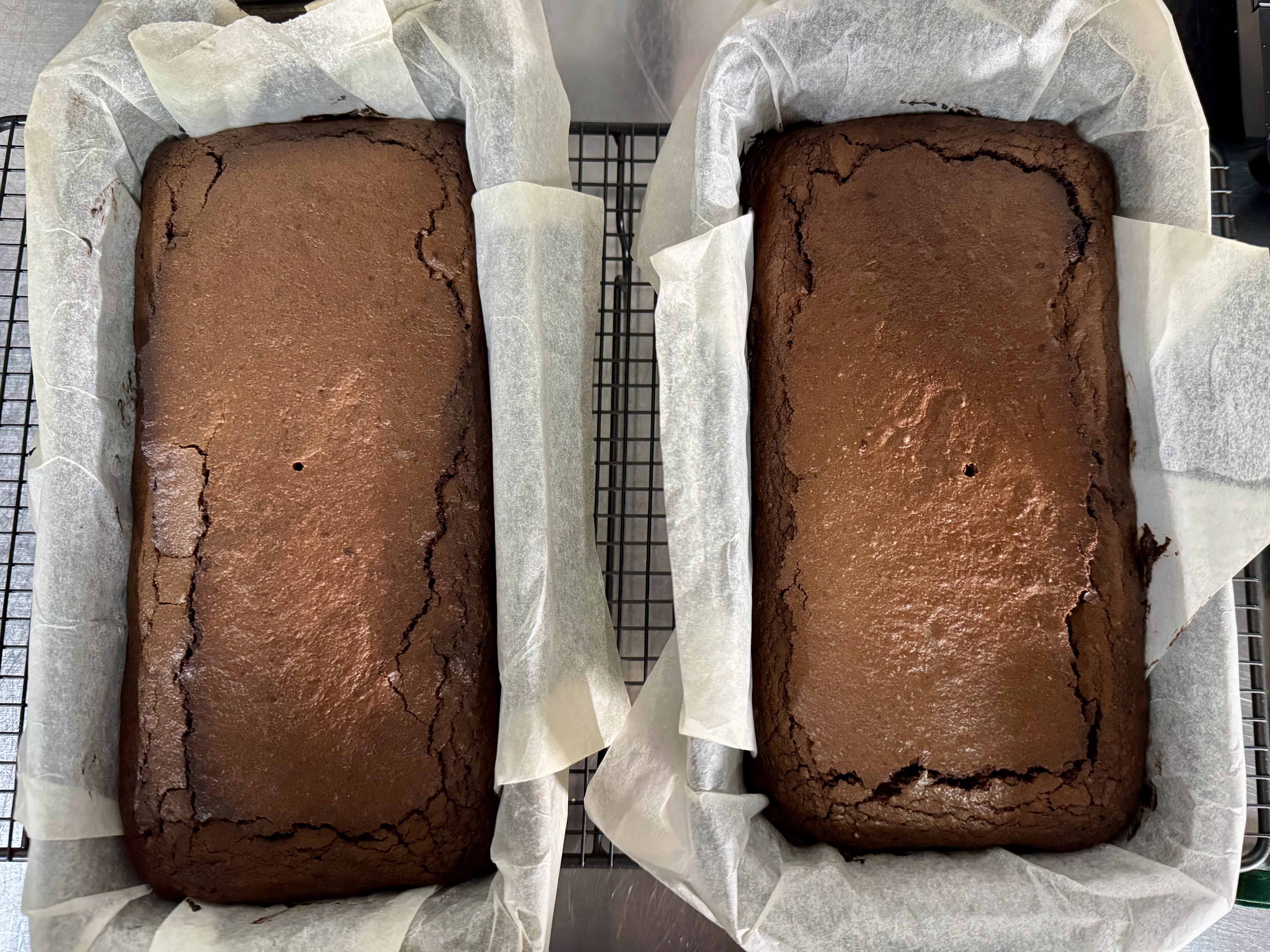 Two rectangular chocolate loaves in parchment-lined tins sit side by side on a wire cooling rack, viewed from above. The tops are glossy with cracked edges and small holes near the center.