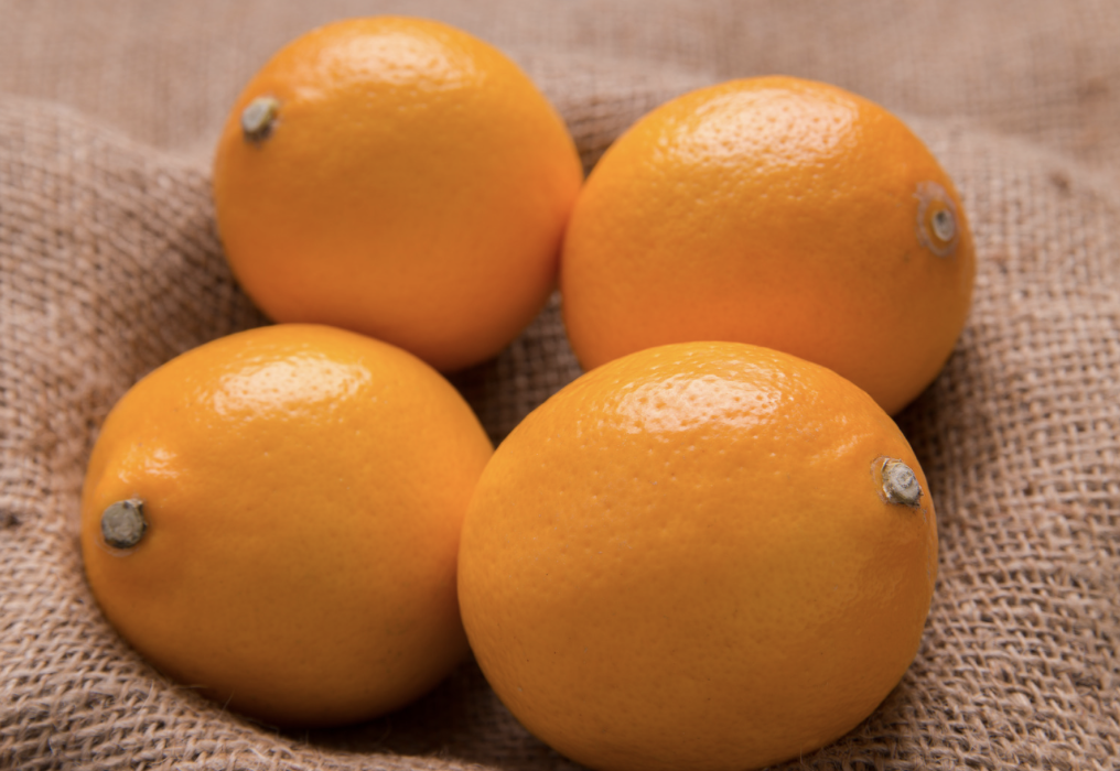 Four smooth, orange-yellow Meyer lemons arranged in a cluster on a burlap fabric surface. The lemons fill most of the frame with their stem ends visible.