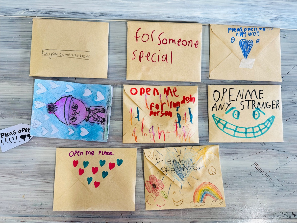 Seven small envelopes and a card are arranged on a gray wooden surface, each decorated with children’s drawings and handwritten messages like “for someone special” and “open me.” The designs include hearts, a blue heart, a smiling face, a rainbow, and a cartoon figure.