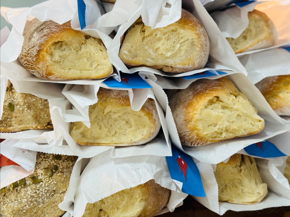 Stacked crusty bread rolls wrapped in white paper sleeves, with a few blue labels visible. Seeded crackers or flatbread pieces sit among the wrapped rolls.