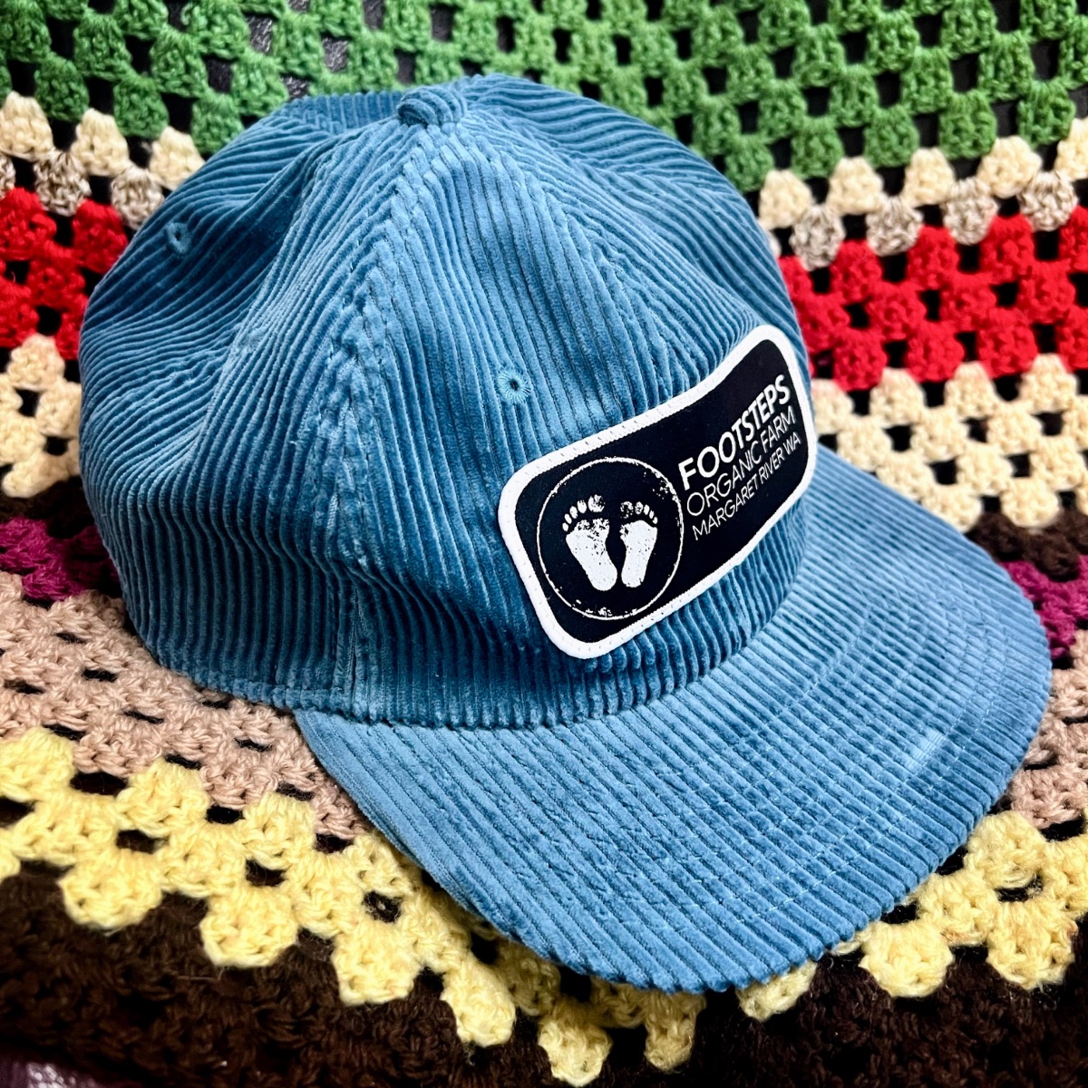 Blue corduroy baseball cap with a black-and-white “Footsteps Organic Farm” patch featuring two footprints. The hat is photographed on a multicolored crocheted blanket.