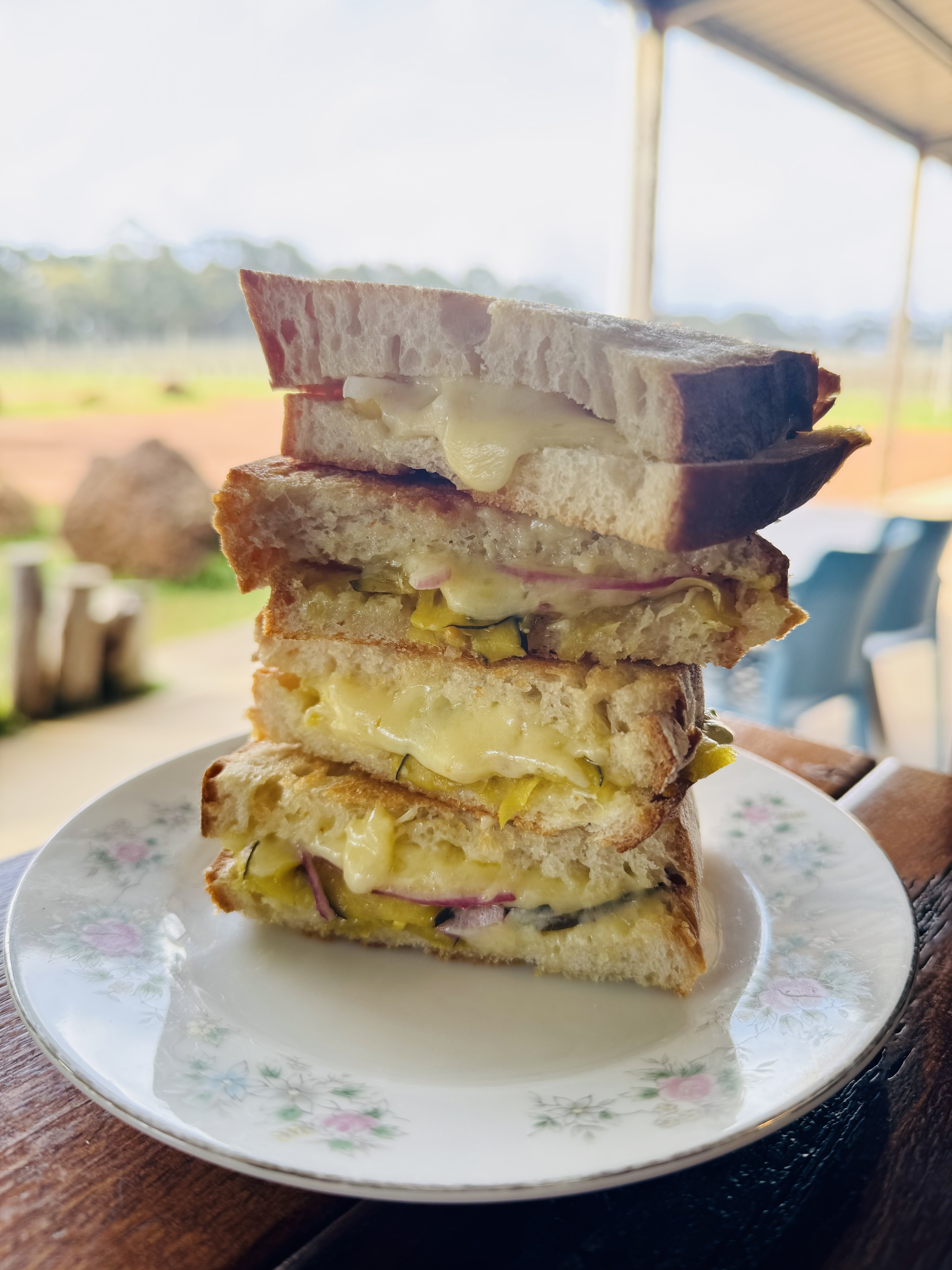 Stack of toasted sandwich halves with melted cheese and pickled vegetables on a floral plate on a wooden table. Blurred outdoor farm view and patio seating in the background.
