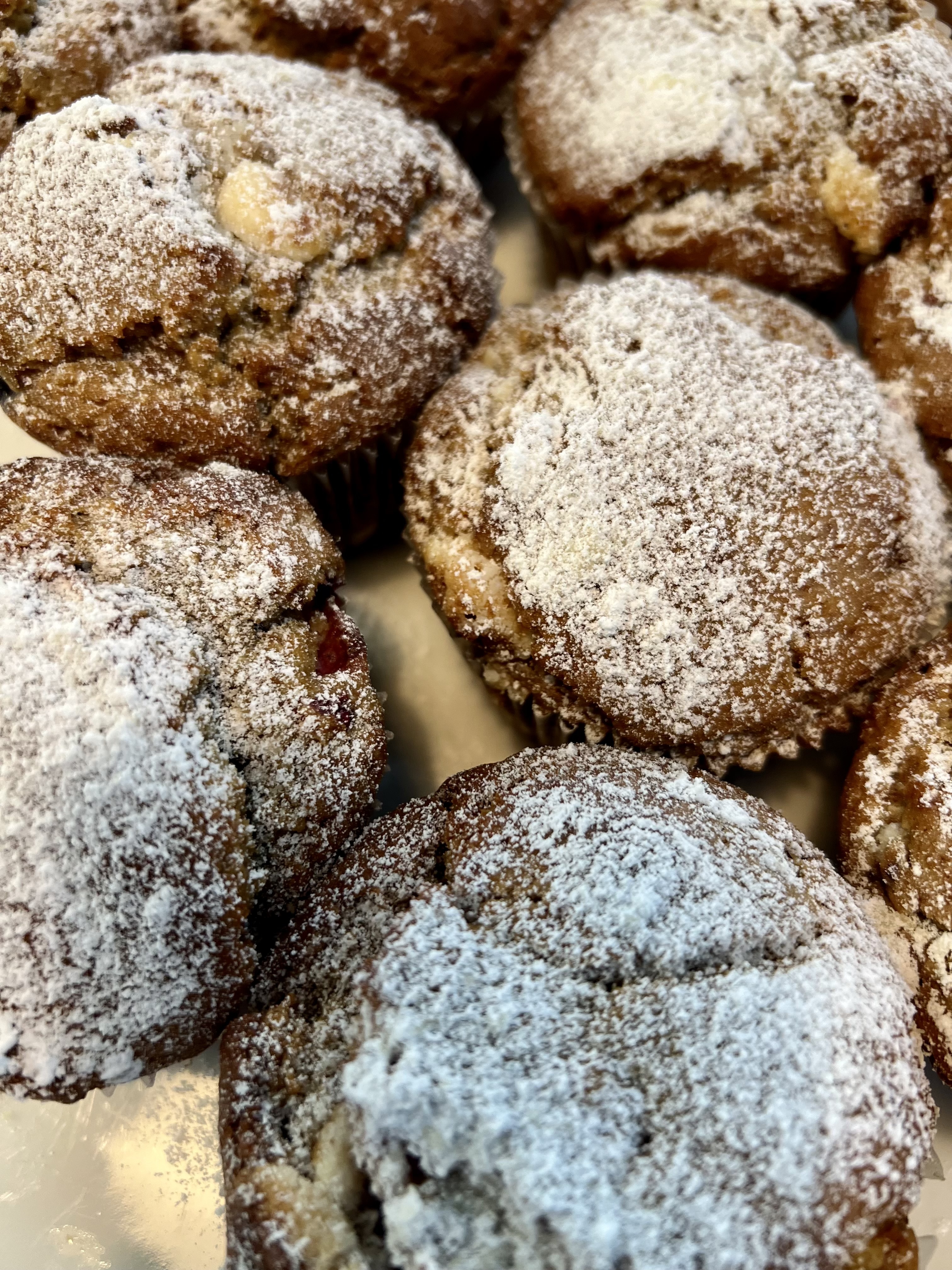 Close-up of several brown muffins on a tray, generously dusted with powdered sugar. Some muffins show bits of fruit or nuts on the surface.