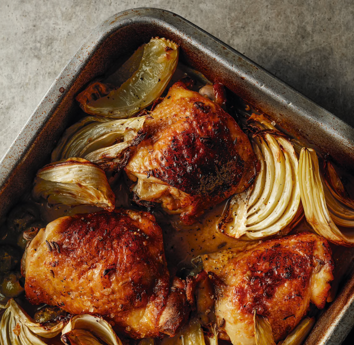 Overhead view of a metal roasting pan with browned chicken pieces and roasted fennel wedges in pan juices. The pan sits on a gray surface with charred edges visible around the rim.
