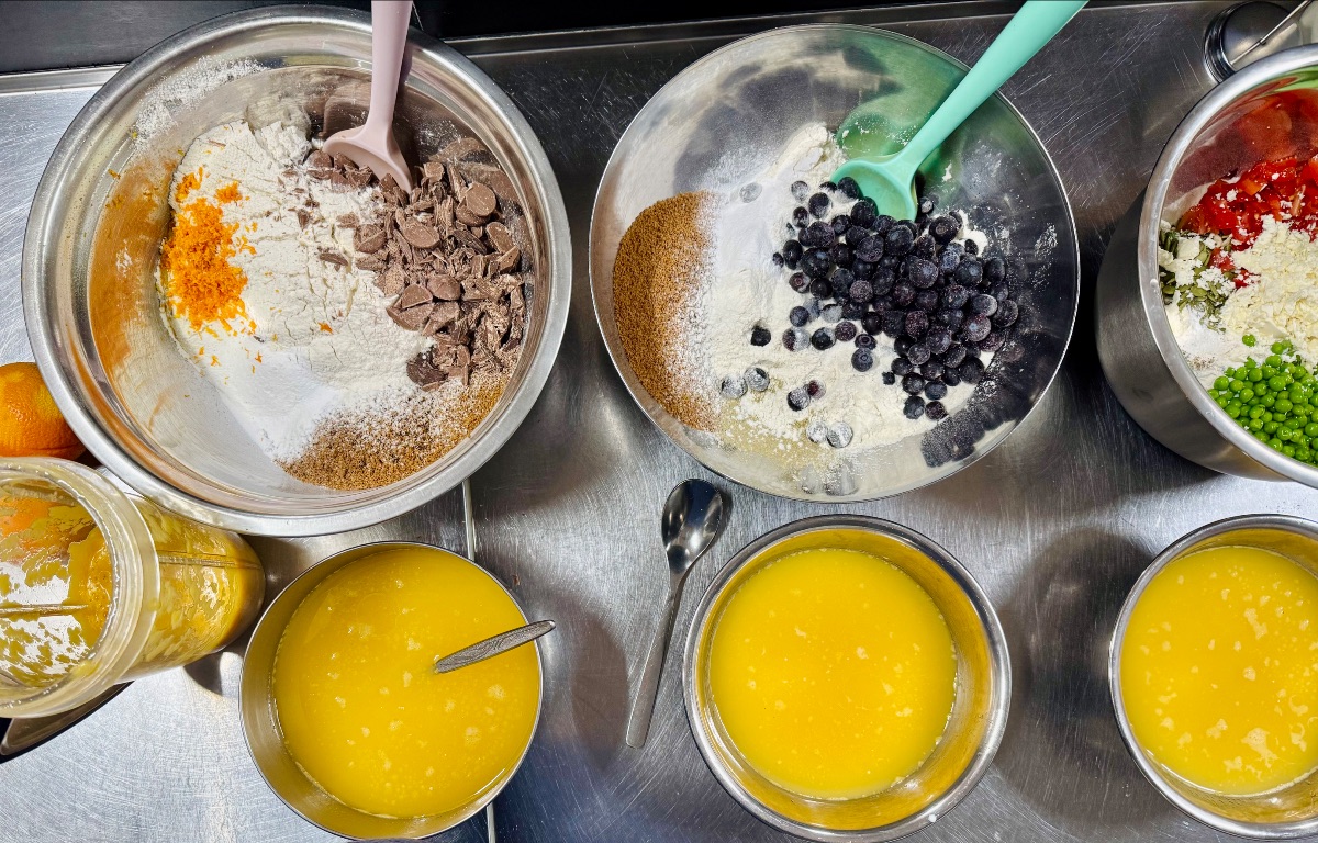 Overhead view of a stainless-steel counter with several metal mixing bowls holding dry ingredients, chocolate pieces, orange zest, and blueberries, each with a spatula. Nearby are bowls of yellow liquid with spoons, a jar of thick yellow mixture, and a bowl of peas and chopped vegetables.