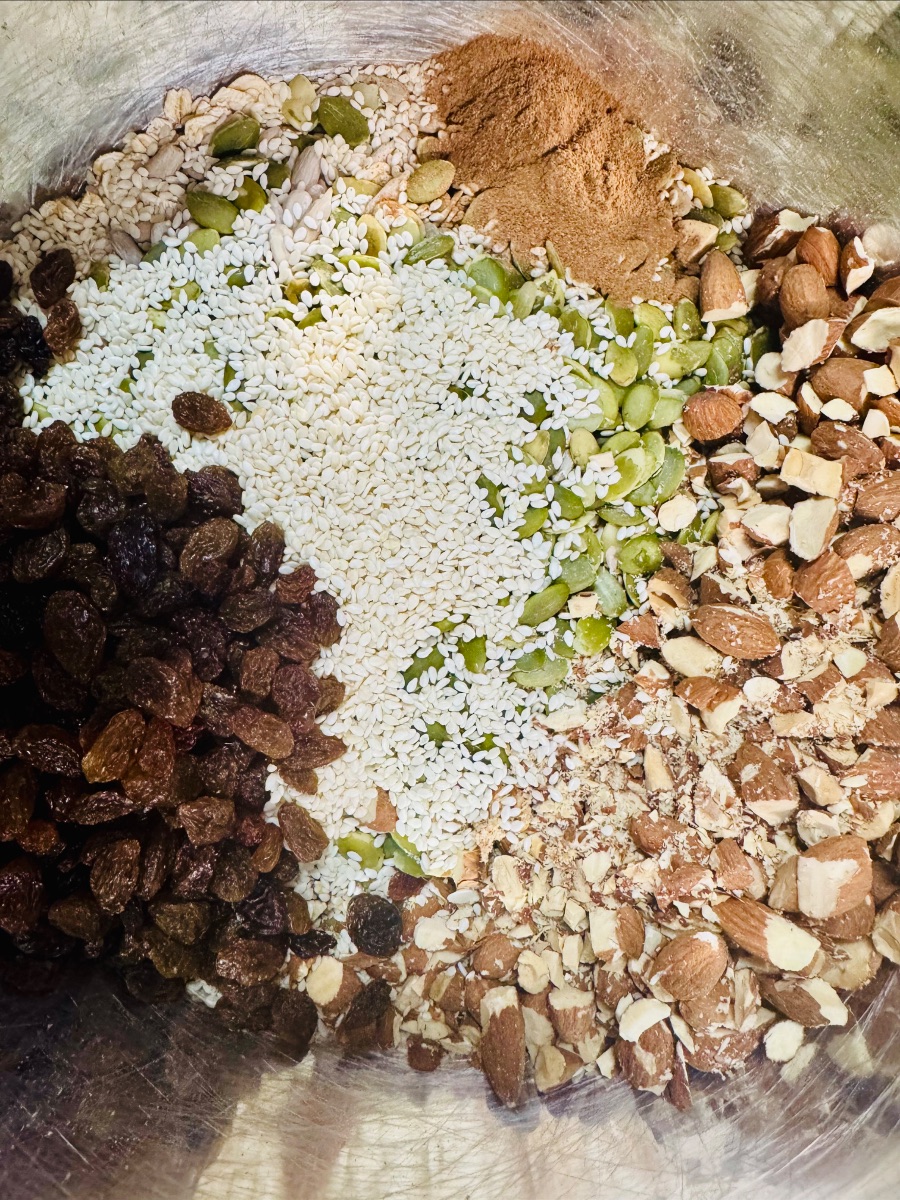 Close-up of a metal mixing bowl filled with separate piles of ingredients: raisins, sesame seeds, pumpkin seeds, chopped almonds, and a mound of brown powder. The ingredients are arranged side by side, not yet mixed.