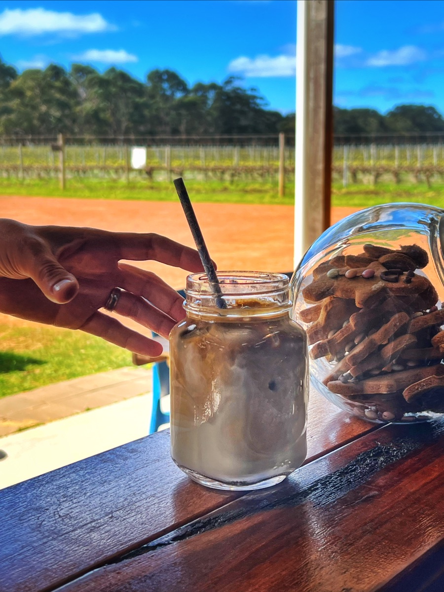 Hand reaching toward a mason jar of iced coffee with a straw on a wooden counter. Beside it is a glass dome filled with biscuits, with a vineyard and blue sky in the background.