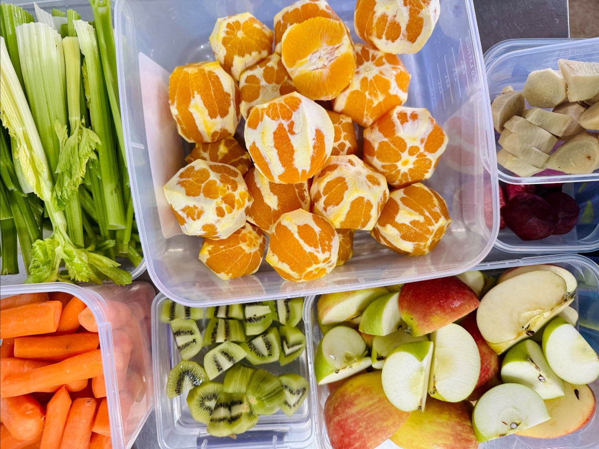 Overhead view of clear plastic tubs filled with prepared produce: peeled small oranges, celery stalks, carrot sticks, sliced kiwi, and apple wedges. Smaller tubs on the right contain chopped banana pieces and two small dark red fruits.