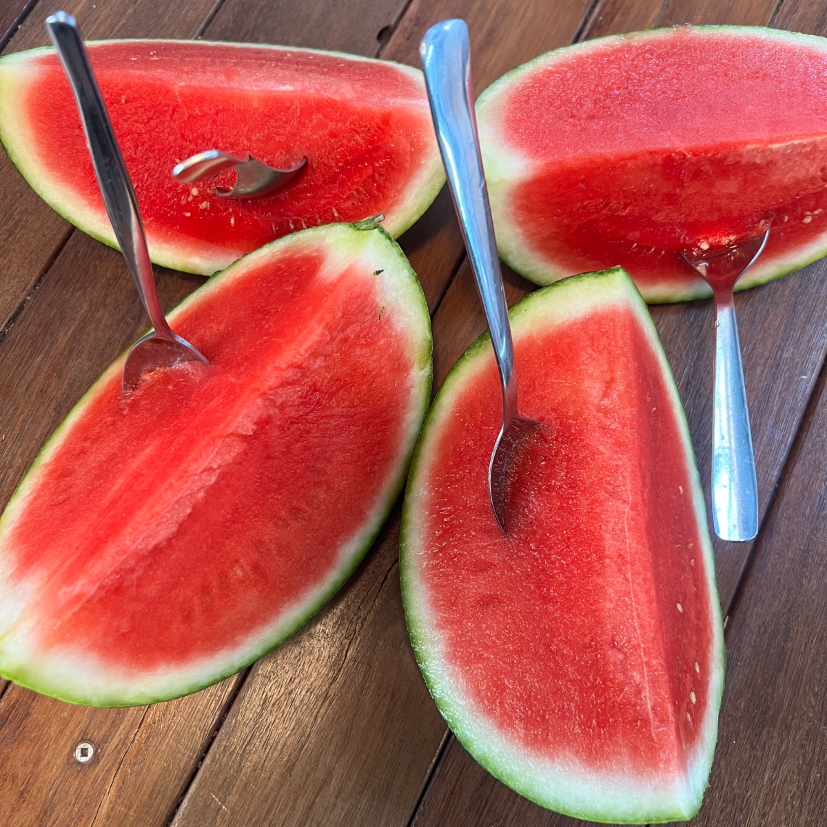 Four watermelon halves sit on a wooden table, each with a metal spoon stuck into the red flesh. The cut rinds are green and white, and the spoons are positioned for scooping.