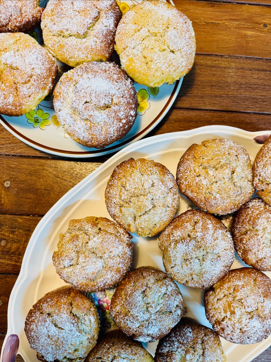 Two plates of golden muffins dusted with powdered sugar sit on a wooden table. One plate is floral-patterned and the other is a white platter with a floral center.