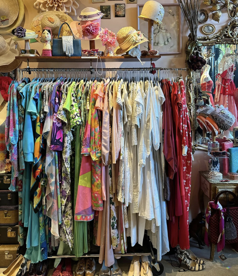 A packed clothing rack holds colorful dresses, blouses, and patterned garments on hangers. Above it, a shelf displays vintage-style hats, purses, gloves, and flowers beside an ornate mirror and framed wall art.