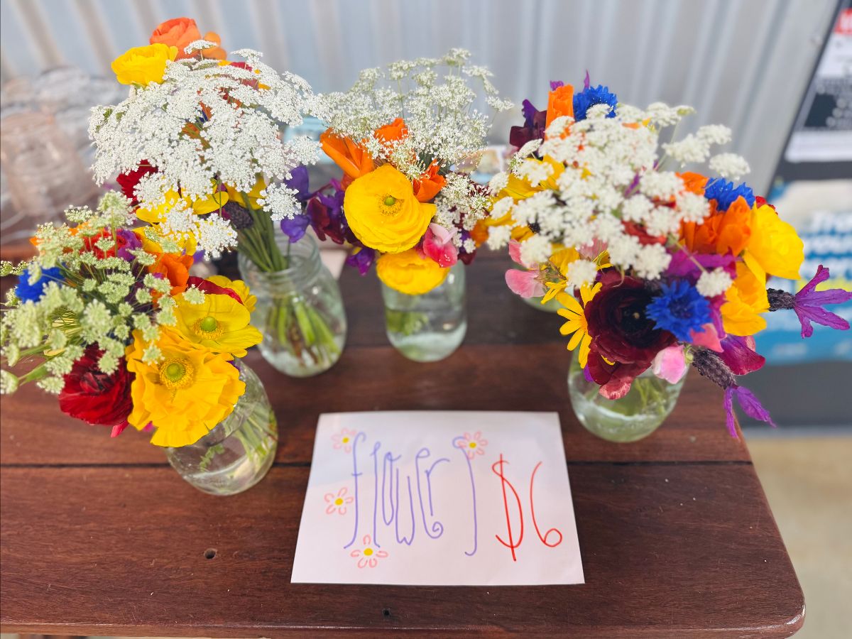 Three small bouquets of colorful flowers in clear glass jars sit on a wooden table. A handwritten sign in front reads “Flower $6.”