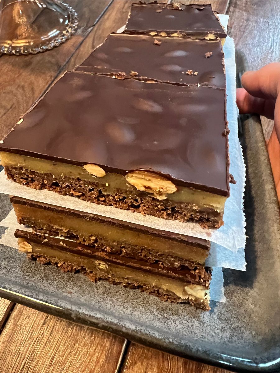 Stack of layered chocolate slice bars with a glossy dark chocolate top, separated by parchment paper, on a tray. A hand is visible at the right edge beside the stack.