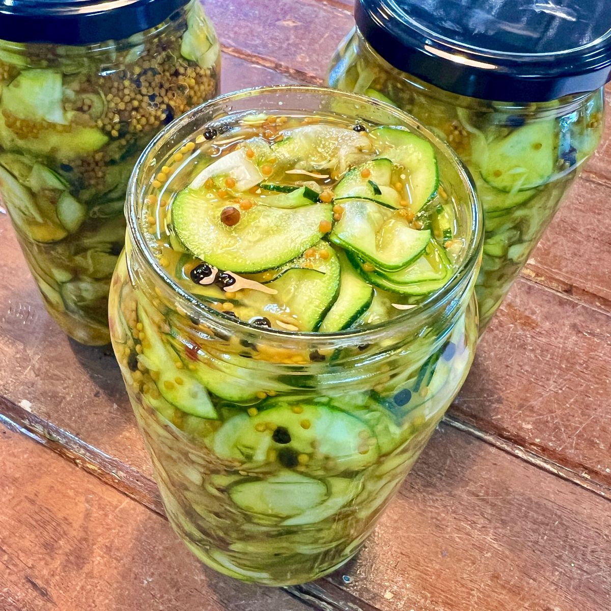 Three glass jars of sliced cucumber pickles in brine sit on a wooden table, with mustard seeds and peppercorns visible. The front jar is open, and two jars behind it have black lids.