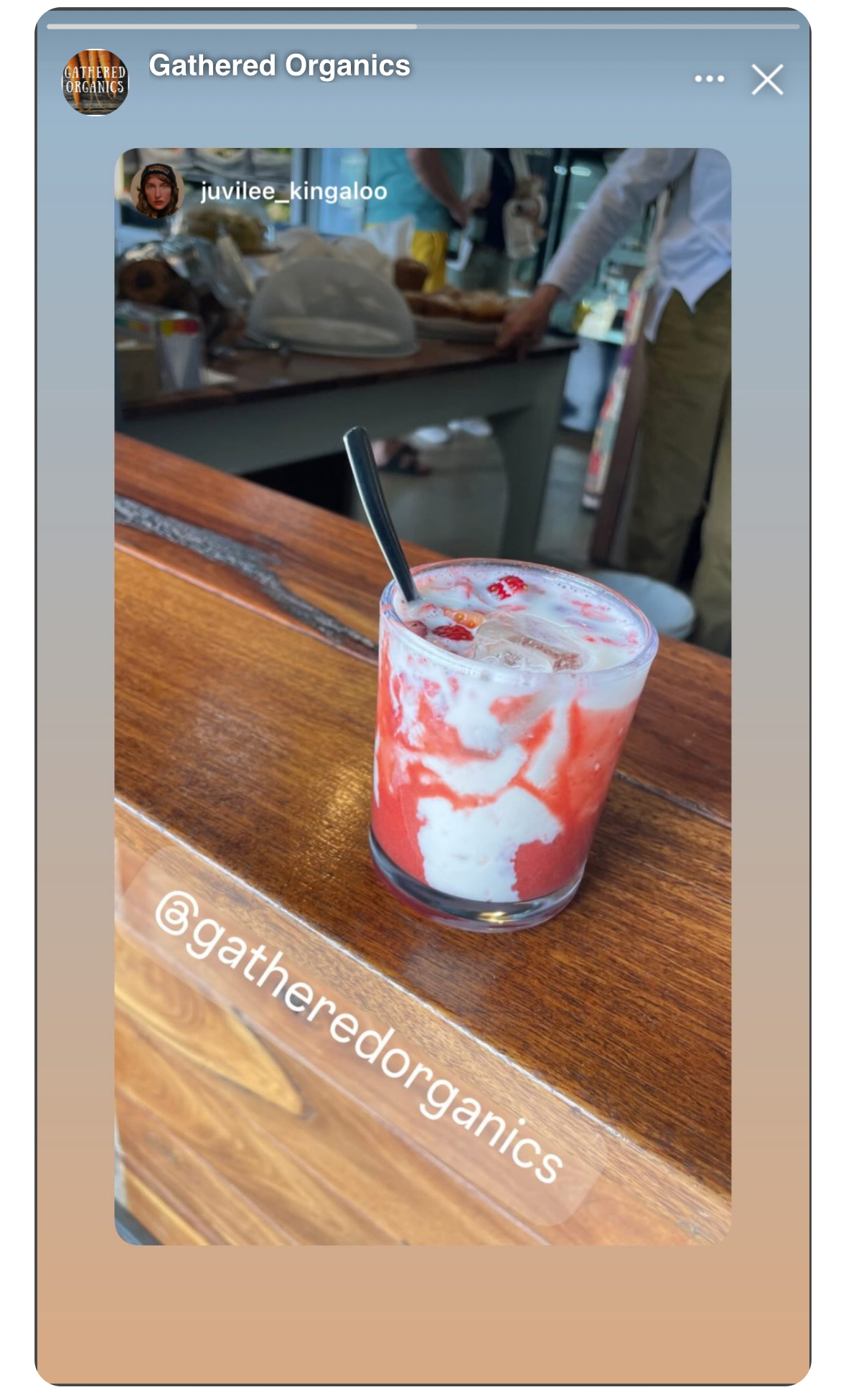 A glass of iced pink-and-white drink with visible strawberry pieces and a straw sits on a wooden counter. In the blurred background are people and a table with food items, with “Gathered Organics” shown at the top.
