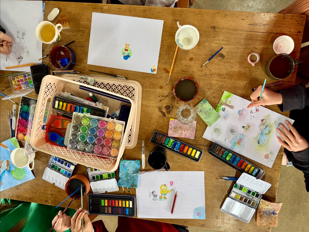 Overhead view of a wooden table covered with watercolor paint sets, brushes, cups, and a basket of art supplies. Children’s hands are painting and drawing on several sheets of paper with colorful sketches.