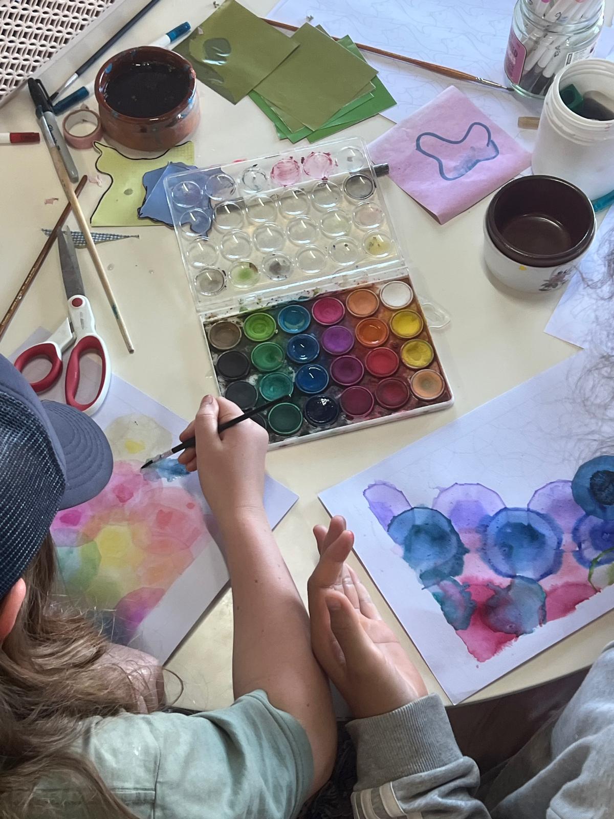 Two children sit at a table painting with watercolors, with a paint palette, brushes, and scissors scattered around. Papers on the table show colorful watercolor blobs and a face-like shape with large blue circles.