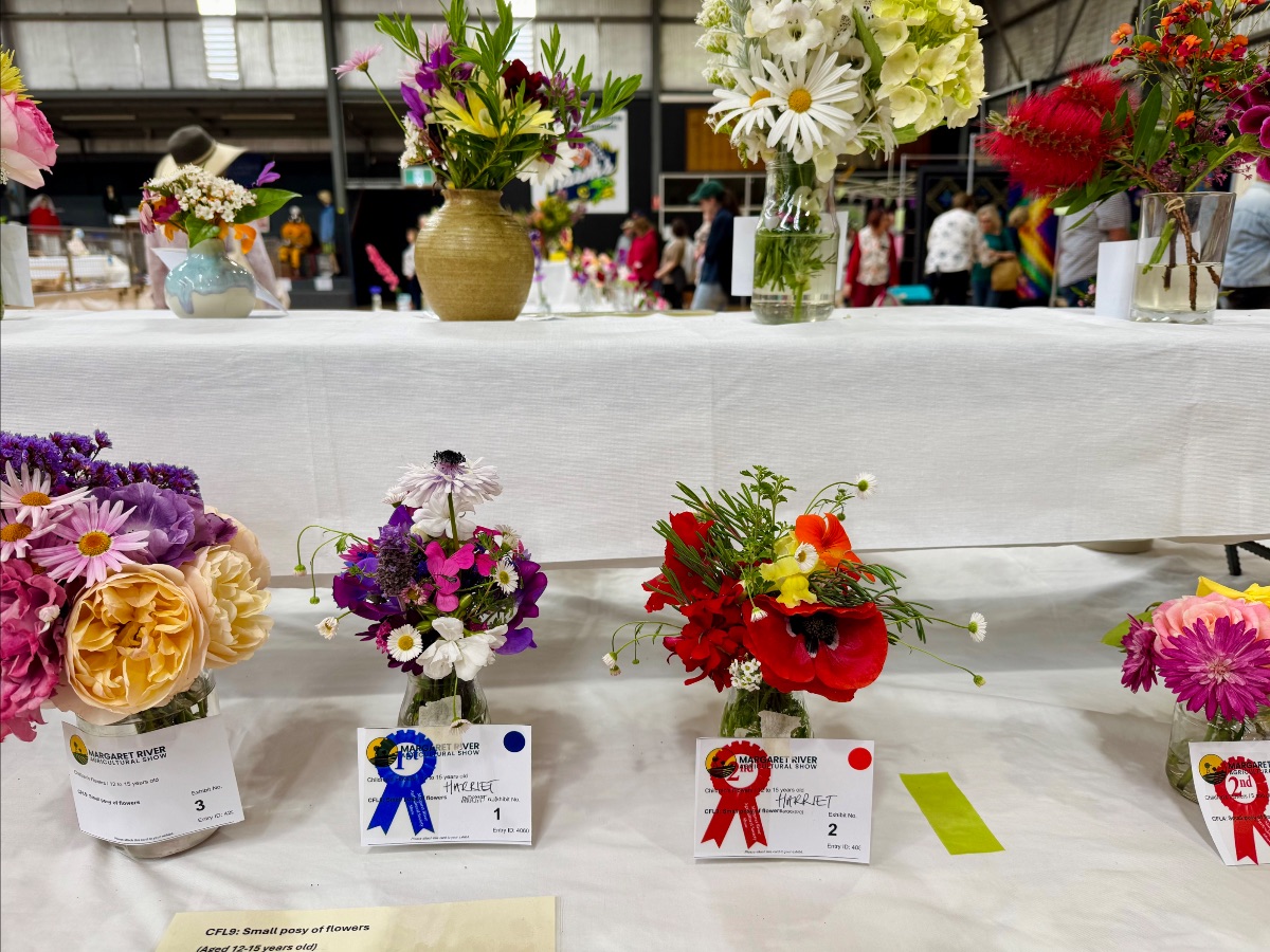 Prize-winning flower posies in small glass jars sit on white-draped tiered tables, each with ribbons and entry cards. In the background, people walk around inside a large indoor hall with other floral displays.