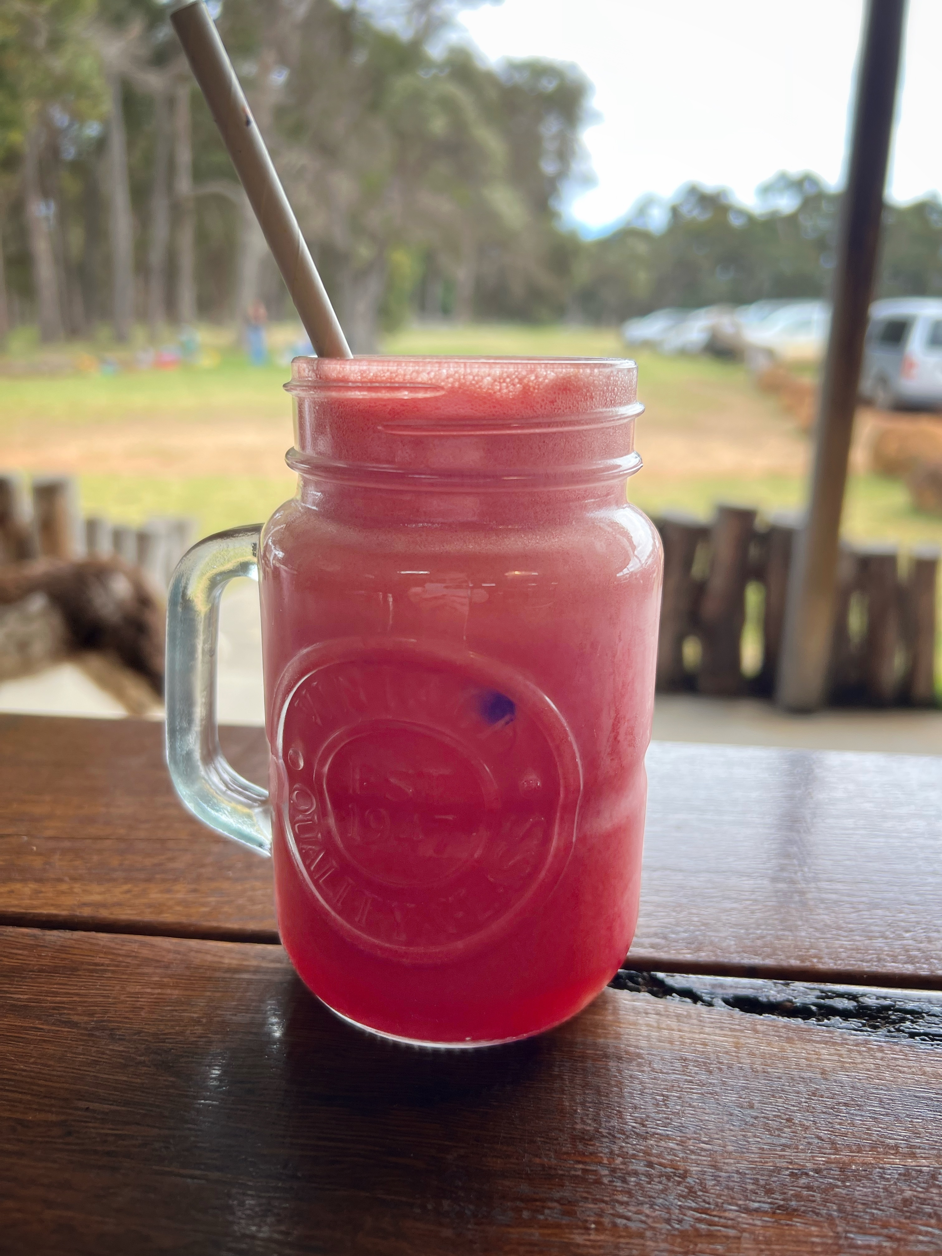 A mason jar mug filled with a bright pink iced drink sits on a wooden table with a paper straw. In the blurred background are trees, a field, and parked cars.