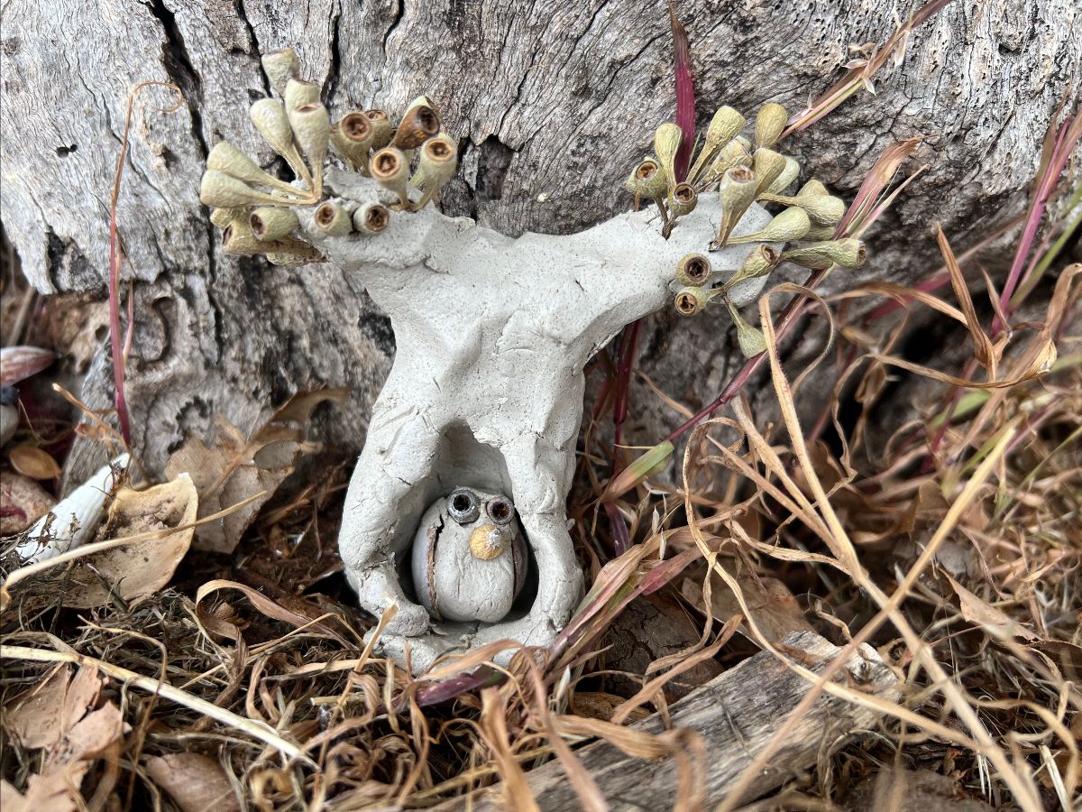 Small clay sculpture of a tree with branch-like arms decorated with dried seed pods, placed against a rough tree trunk. A round clay figure with painted eyes sits in a hollow at the base amid dry grass and leaves.
