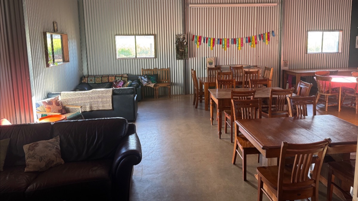 Interior of a corrugated-metal room with wooden tables and chairs on the right and two dark leather sofas with pillows and a throw on the left. Two windows and a small mirror are on the walls, and a string of colorful hanging flags decorates the back wall.
