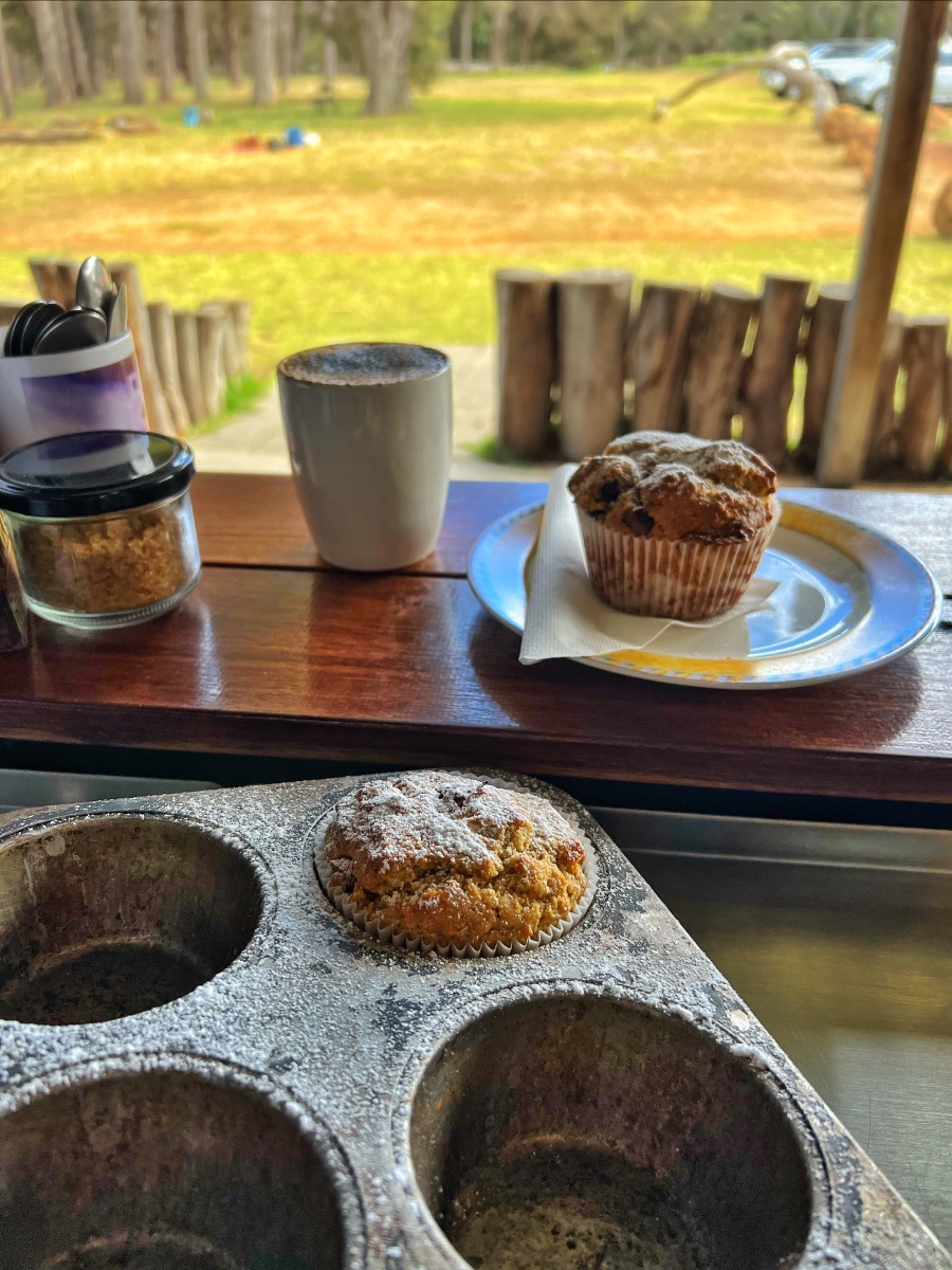 A muffin sits in a worn metal muffin tin in the foreground. On a wooden counter behind it are a mug and another muffin on a plate, with a grassy outdoor area and rustic fence in the background.