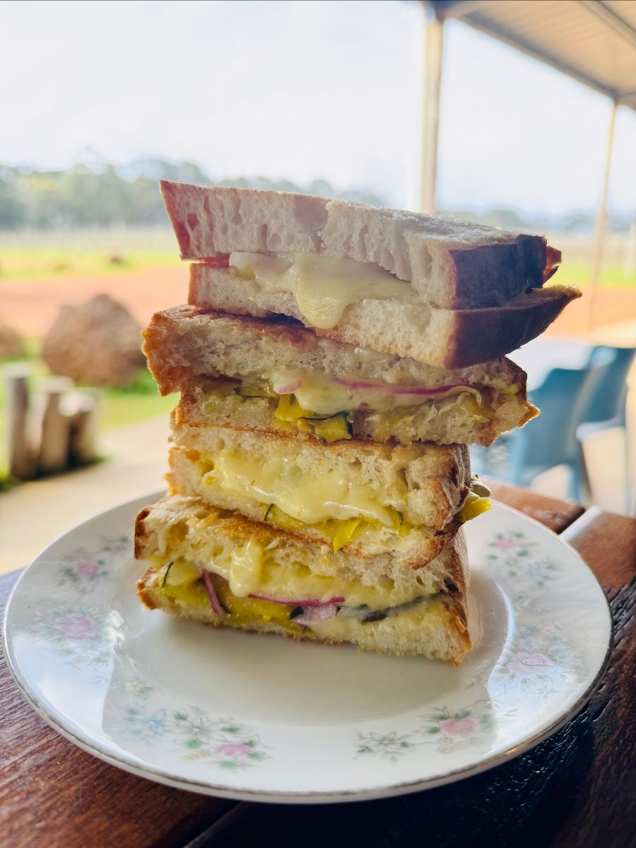 Stack of toasted sandwich quarters with melted cheese and sliced vegetables on a floral plate. The plate sits on a wooden table with a blurred outdoor farm view in the background.