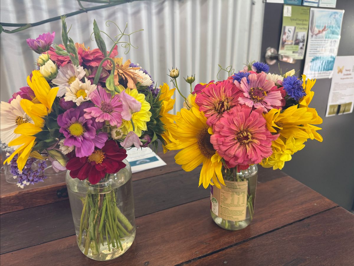Two mixed bouquets of bright flowers—pink and purple daisies, yellow sunflowers, and other blooms—sit in clear glass jars on a wooden table. A corrugated metal wall and a bulletin board with flyers are in the background.