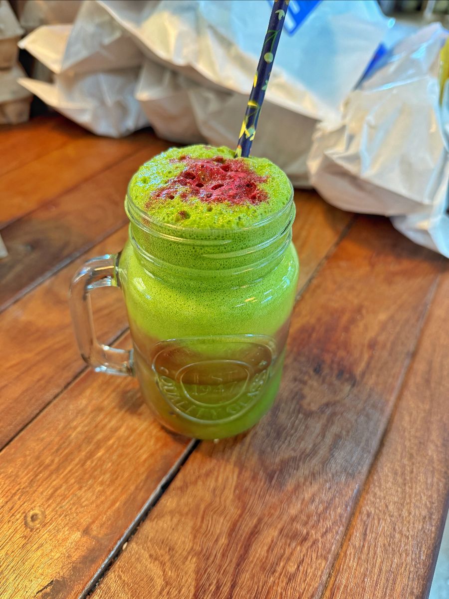 A glass mason-jar mug filled with a frothy green drink sits on a wooden table, topped with a red sprinkle and a dark straw. White paper-wrapped packages are blurred in the background.