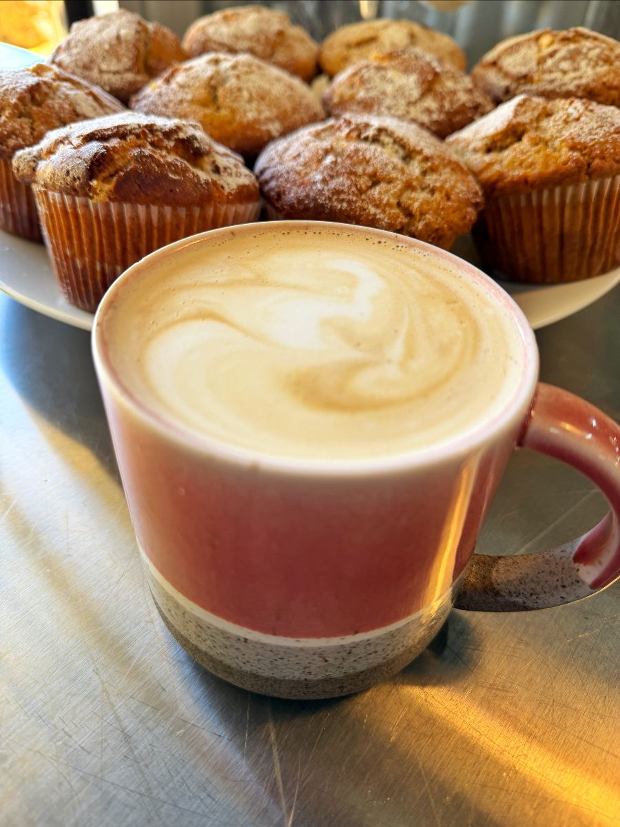 A pink ceramic mug of foamy coffee sits on a stainless-steel counter. Behind it, a plate holds several sugar-dusted muffins in paper liners.
