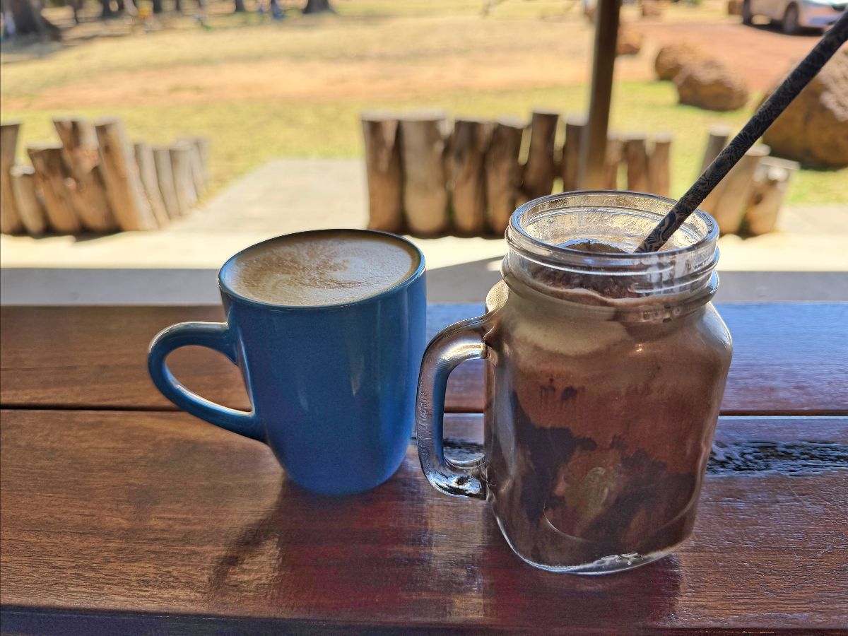 Blue mug of coffee and a handled glass jar of iced drink with a straw sit on a wooden table. In the blurred background are log stools and a grassy outdoor area.
