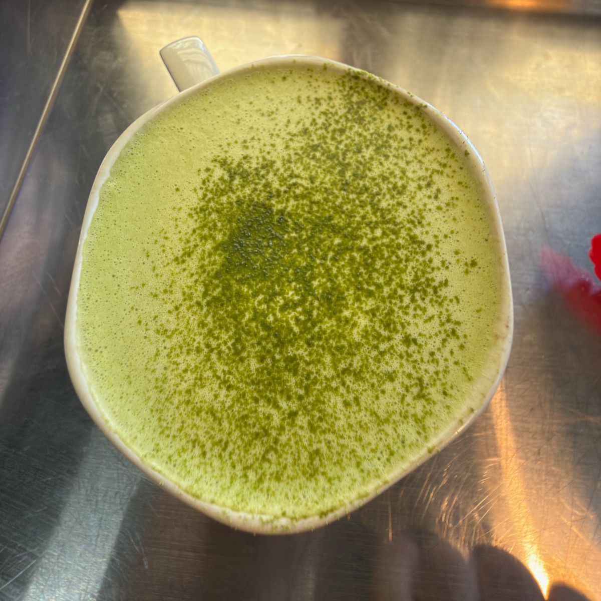 Overhead view of a white mug filled with a pale green foamy drink, dusted with darker green powder. The mug sits on a shiny metal countertop.