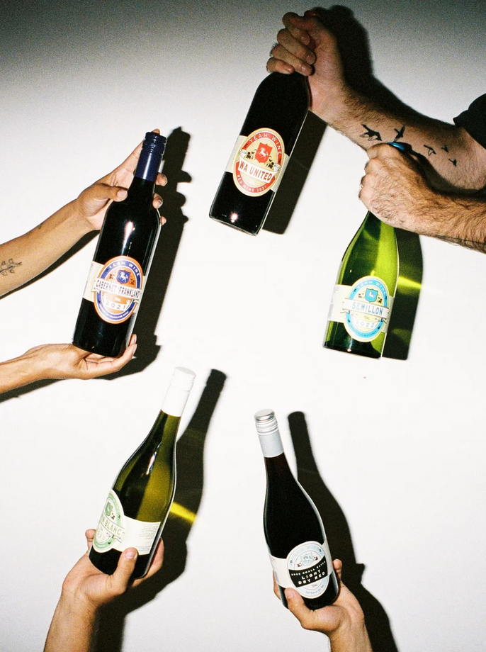 Five hands hold up assorted Yeah Wine bottles against a plain light background, including dark red and green glass with colourful round labels