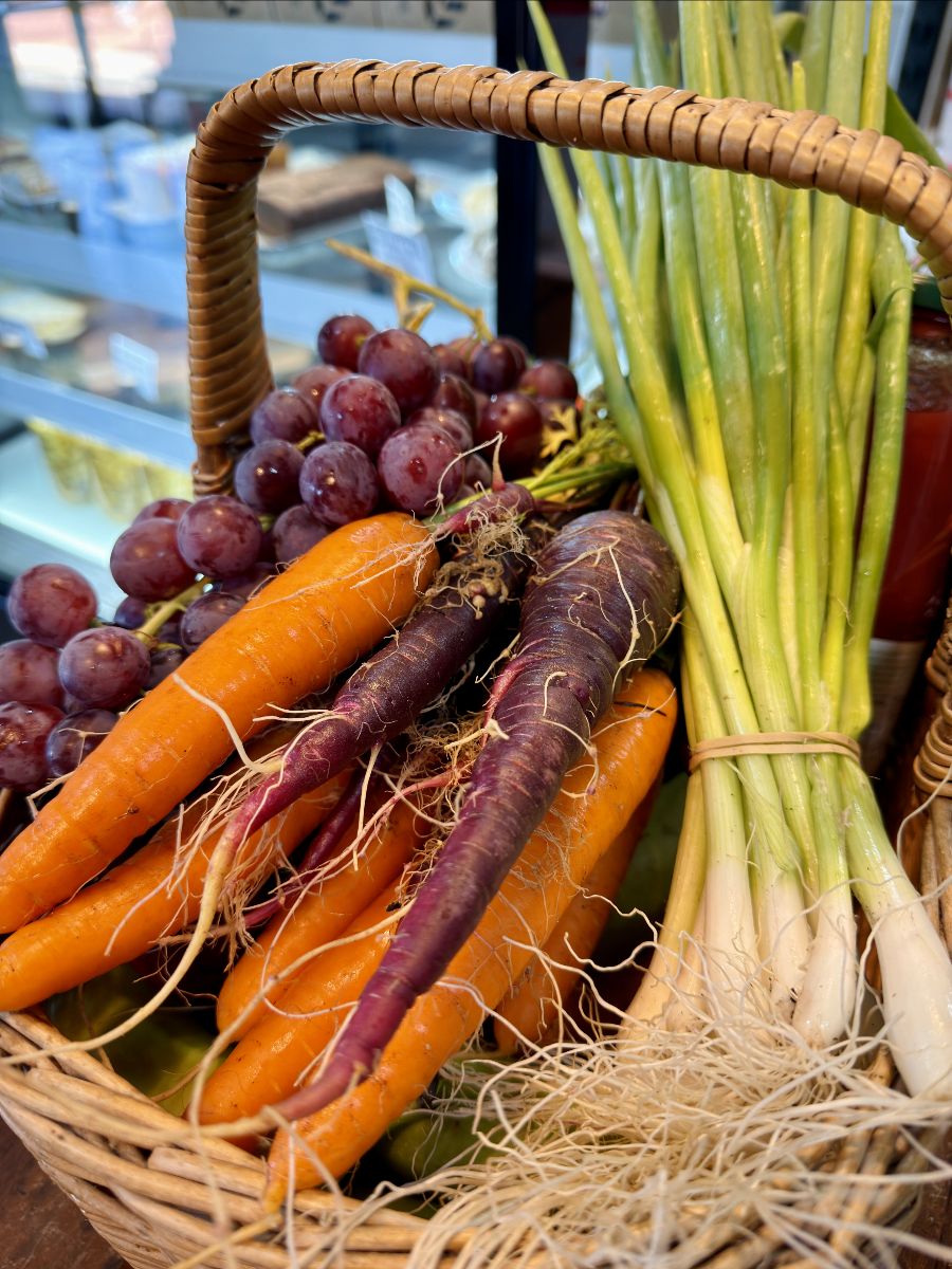A wicker basket filled with orange and purple carrots with roots, a bunch of spring onions, and a cluster of red grapes, displayed in the farm shop