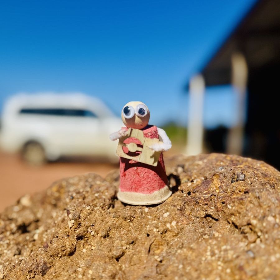 A small handmade figurine with googly eyes and a pink dress perched on a rock outdoors with a blue sky and farm buildings in the background