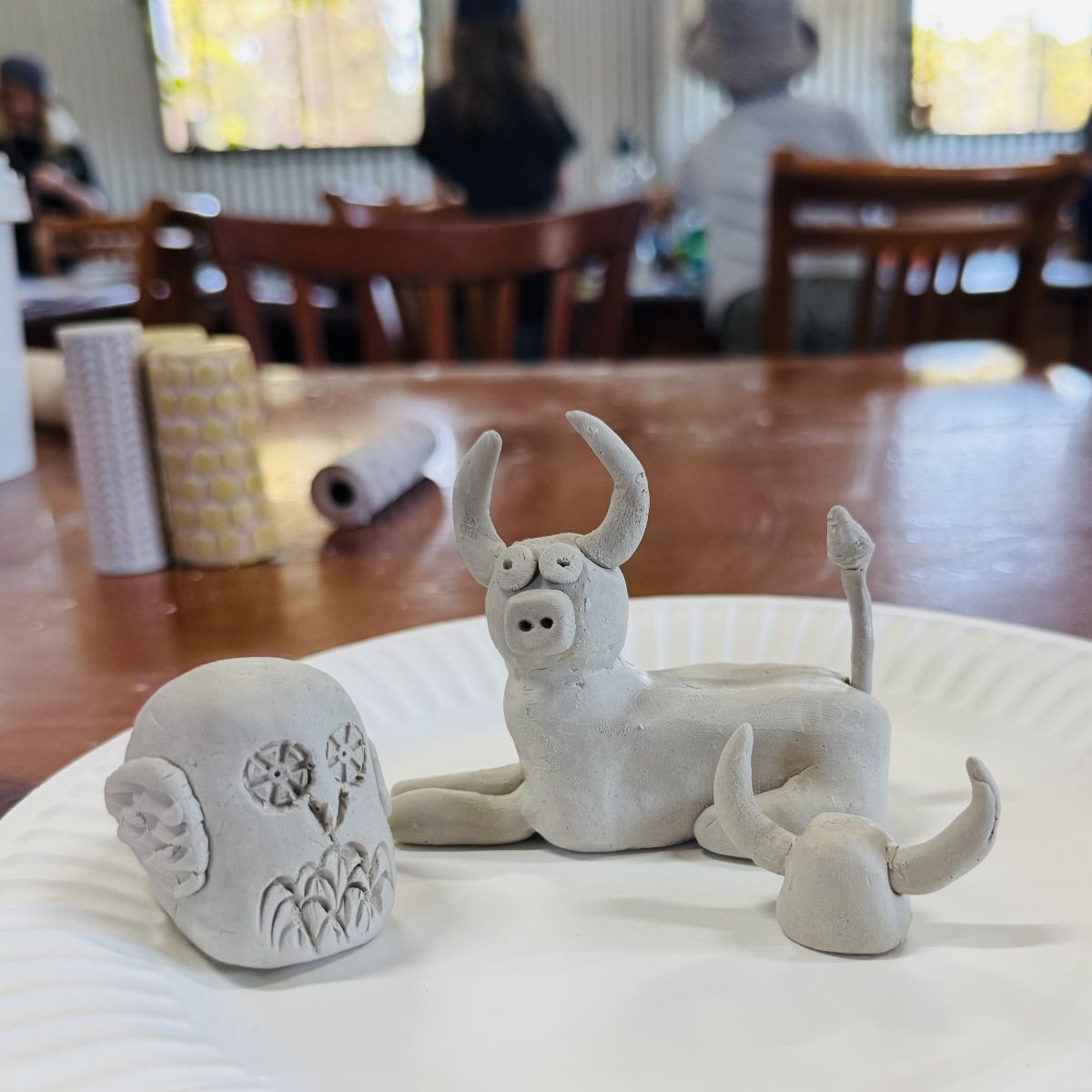 Handmade clay sculptures on a paper plate including a decorated owl-shaped piece with flower carvings and a horned animal figure, made by homeschool kids at the farm