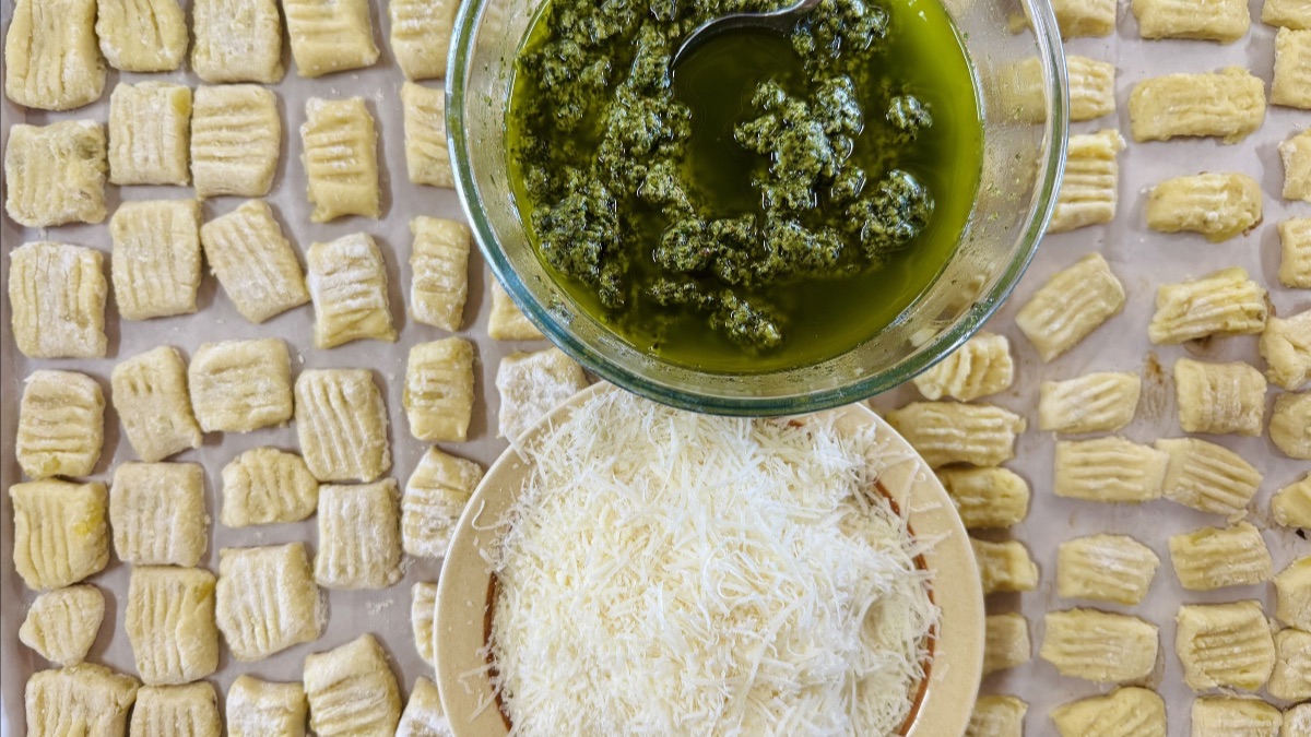 Rows of handmade potato gnocchi laid out on baking paper with a glass bowl of basil pesto and a plate of grated parmesan cheese in the centre