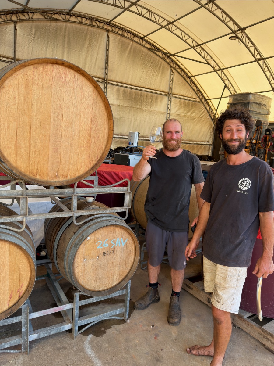 Two men stand inside a large arched shed beside stacked wooden barrels on metal racks, with one holding up an empty wine glass. Machinery and covered containers are visible in the background.