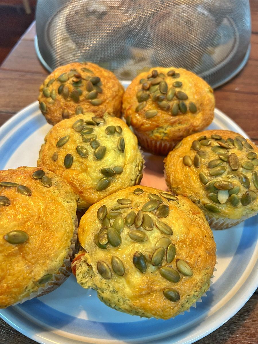 Six golden muffins topped with pumpkin seeds sit on a white plate on a wooden table. A mesh food cover is visible in the background.