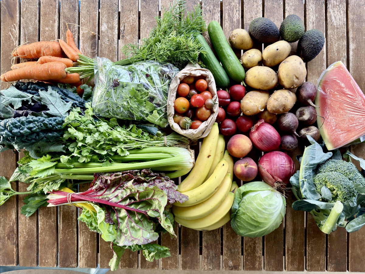 Assorted fresh produce arranged on a wooden table, including carrots, leafy greens, celery, cucumbers, potatoes, avocados, bananas, tomatoes, plums, red onions, beets, cabbage, broccoli, and a wrapped wedge of watermelon. A paper bag of cherry tomatoes sits near the center.